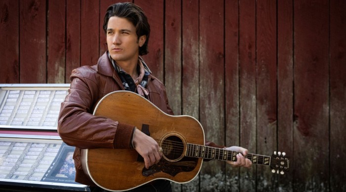 A man wearing a brown leather jacket holding an acoustic guitar